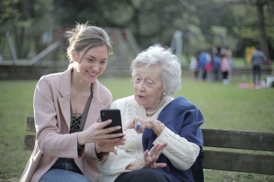 Una señora mayor y una mujer joven sentadas en un banco de un parque, mirando un teléfono juntas y sonriendo.